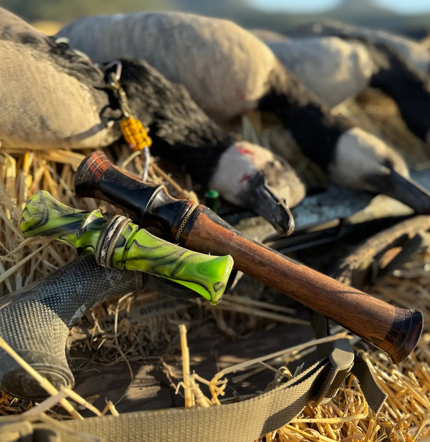 lime green and black swirl design short reed goose call lain next to black and white ebony goose flute with hunt shot of several bagged canada geese and rifle in the background