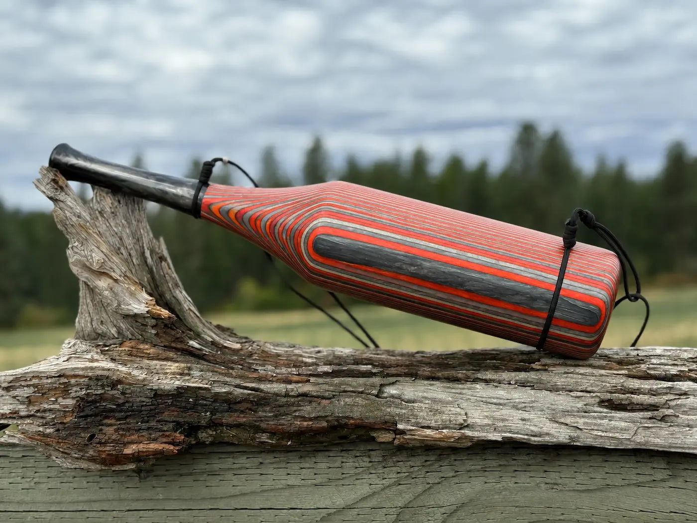 A handmade wooden elk bugle with red, silver, and gray striped pattern, resting on a piece of driftwood outdoors with a blurred forest and cloudy sky in the background.