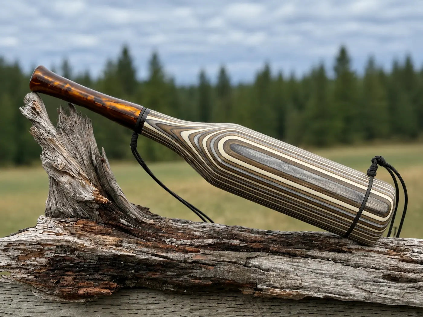 A handmade wooden elk bugle with a natural wood inlay of silver and bronze stripes and a dark bronze mouthpiece, resting on a weathered log with a forest and cloudy sky in the background.
