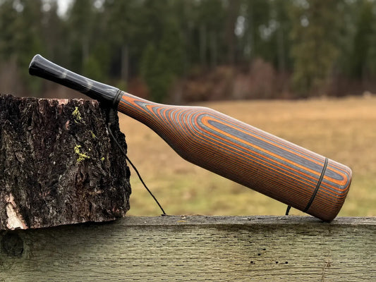 A handmade wooden elk bugle with a smoky black acrylic mouthpiece, resting on a weathered wooden fence next to a tree stump with an outdoor field and forest in the background.