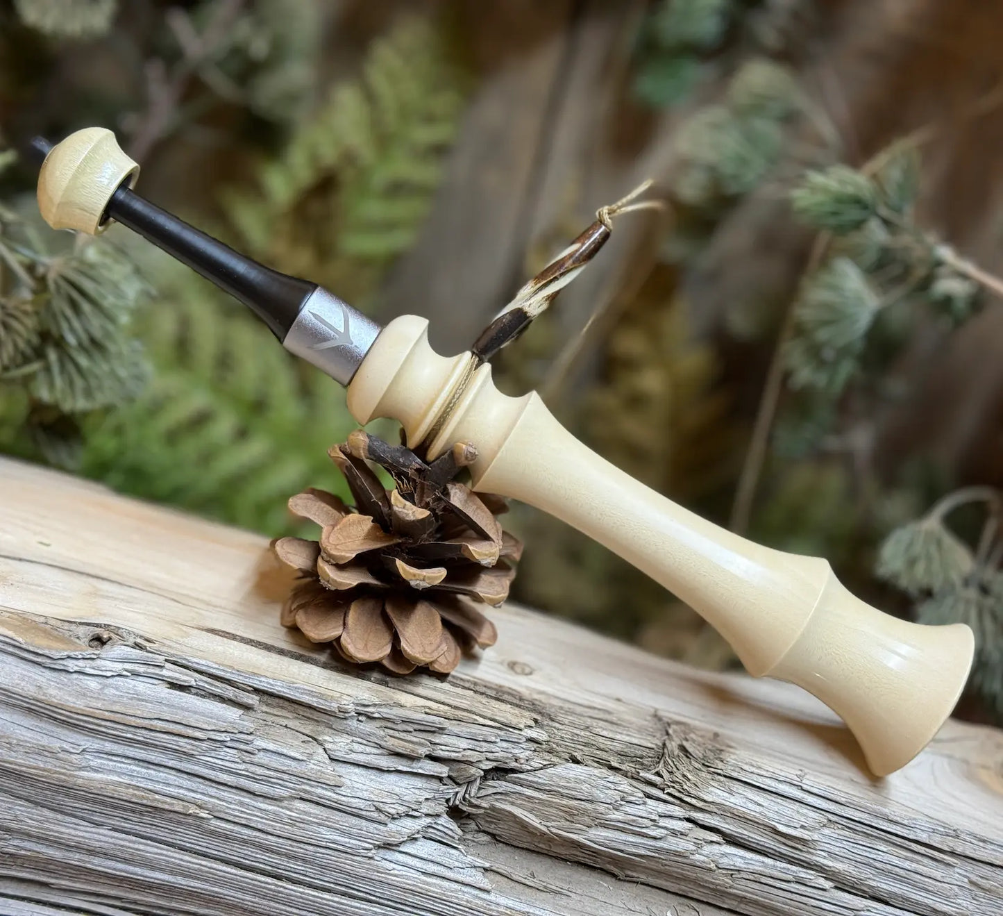 Field Grade Turkey Trumpet in milky light honey tone wood with custom turkey foot band and black tip mouthpiece with matching top of holly wood. The turkey trumpet is leaning on a pine cone for field use or display