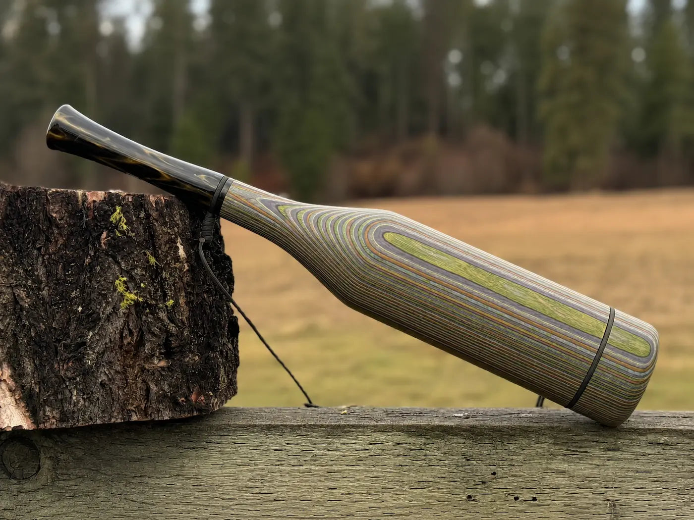 A handmade wood elk bugle with a camo striped grain pattern and smoky venom acrylic mouthpiece, resting on a weathered wooden fence next to a tree stump with a black lanyard strap.