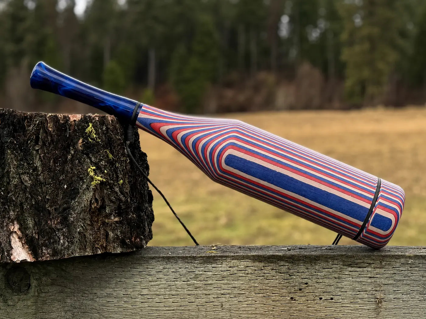 A red and blue striped wooden elk bugle with a blue pearl acrylic mouthpiece resting on a rustic wooden fence next to a tree stump with a blurred forest background.