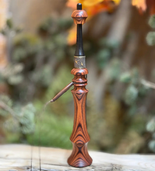 This caramel rosewood turkey trumpet sits upright with an onyx mouthpiece, ornate reddish-brown carved handle, brassy texturized collar with turkey tracks, and a light buffalo horn bead on its lanyard.