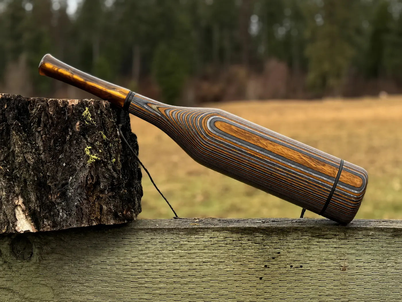 A handmade wooden elk bugle with a dark brown and amber striped pattern, resting on a rustic wooden fence with a tree stump in the foreground and a blurred outdoor field and forest background.