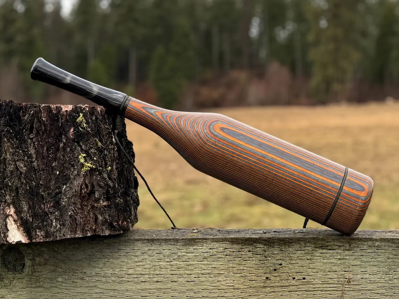 A handmade wooden elk bugle with a smoky black acrylic mouthpiece, resting on a weathered wooden fence next to a tree stump with an outdoor field and forest in the background.