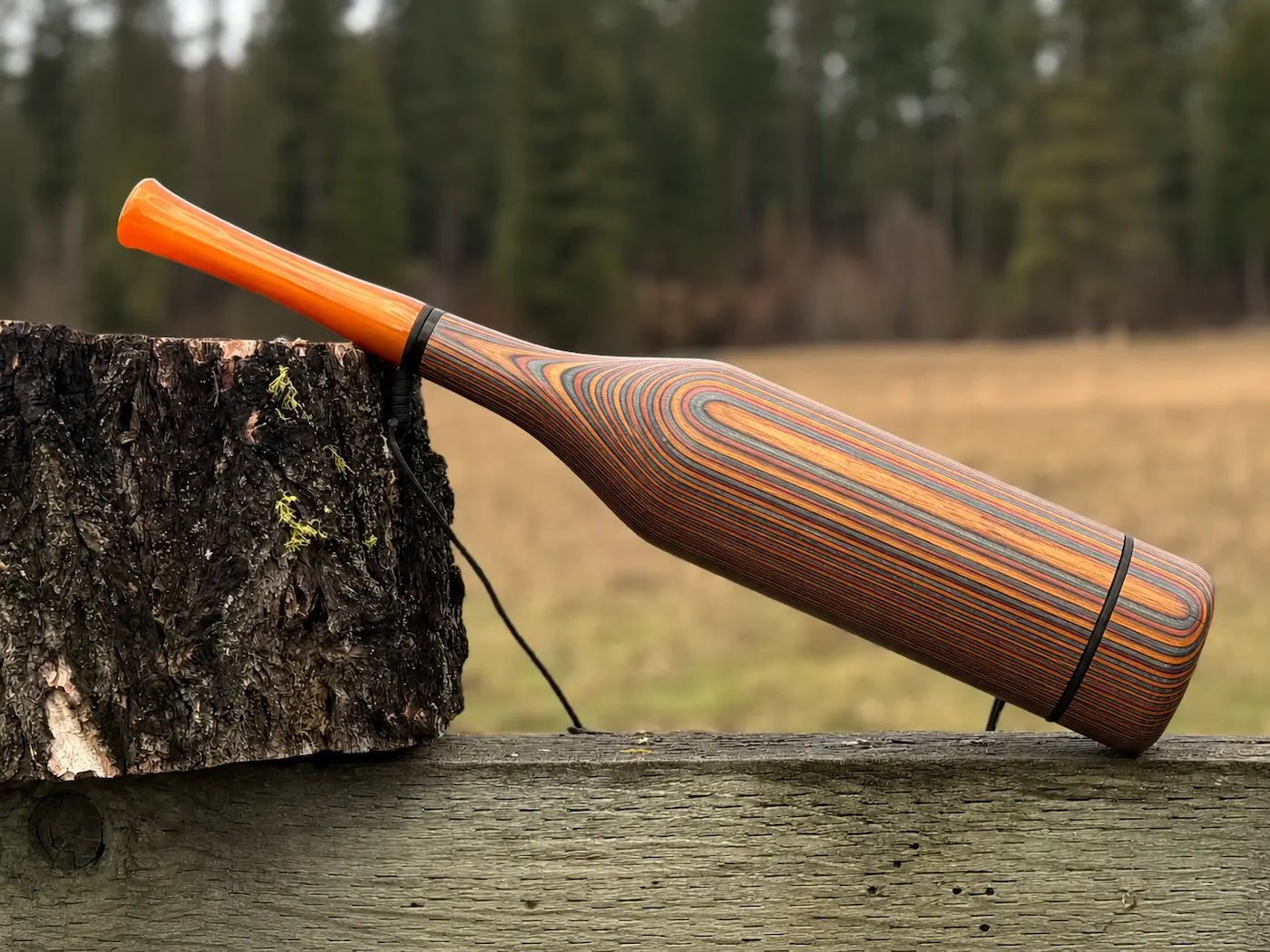 A handmade wooden elk bugle featuring a vibrant orange mouthpiece and a body with a striped wood grain pattern of brown, orange, and silver, resting on a weathered wooden fence with a tree stump and a forest in the background.
