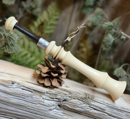 Field Grade Turkey Trumpet in milky light honey tone wood with custom turkey foot band and black tip mouthpiece with matching top of holly wood. The turkey trumpet is leaning on a pine cone for field use or display