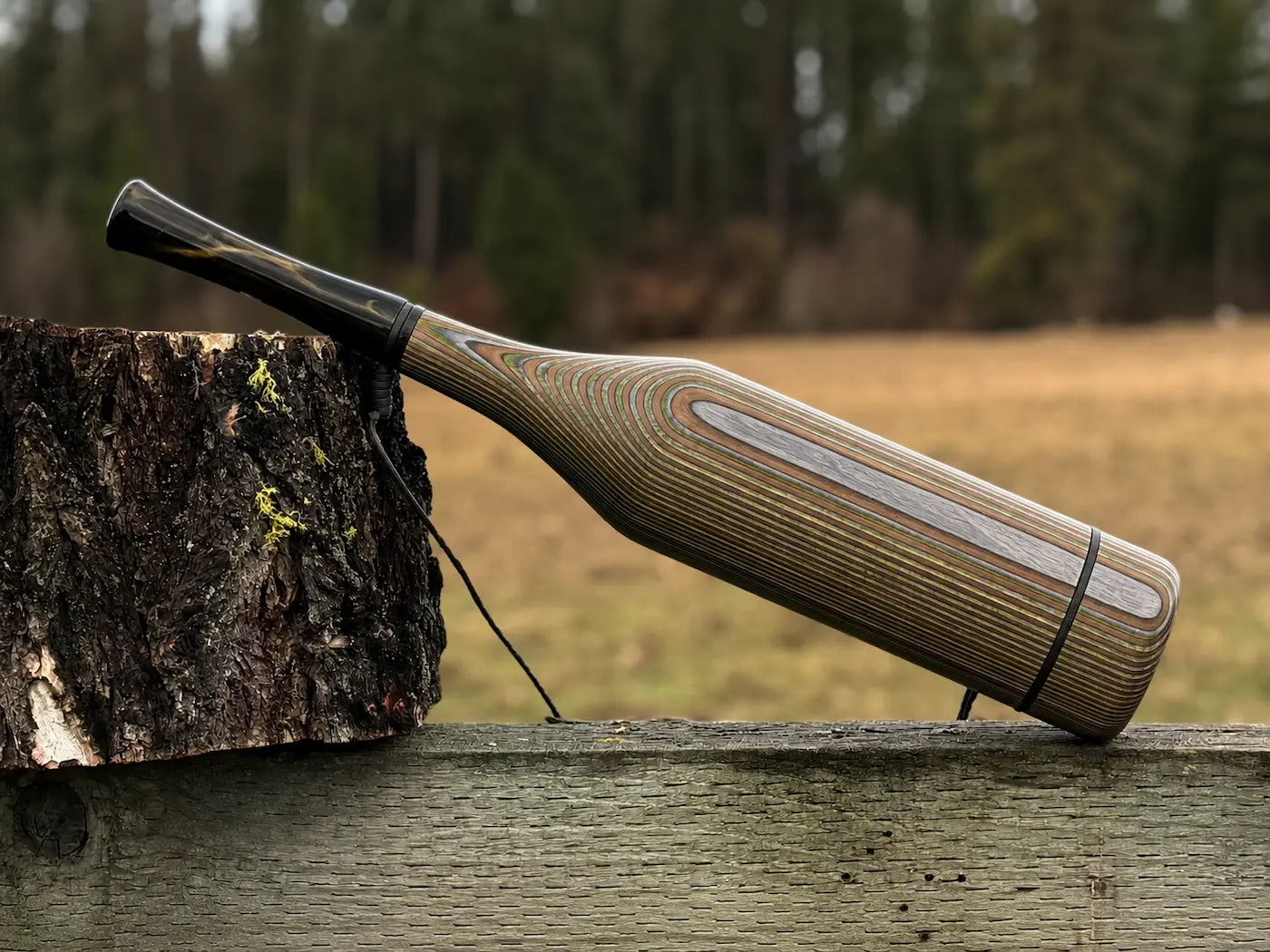 A handmade wood elk bugle with a camo striped grain pattern and smoky black mouthpiece, resting on a weathered wooden fence next to a tree stump with a black lanyard strap.