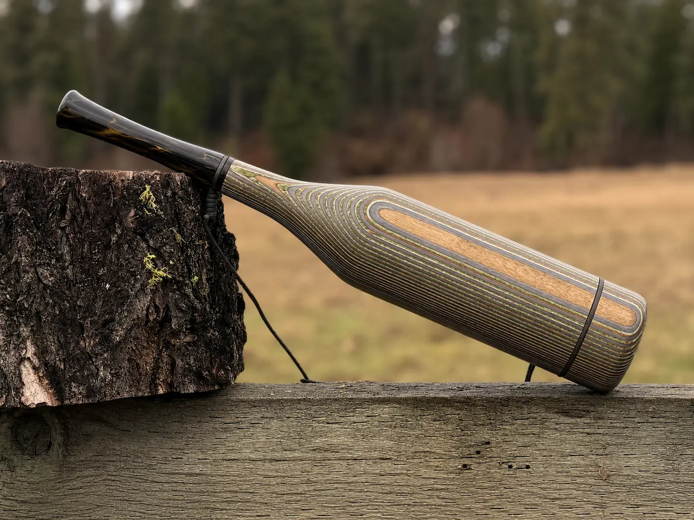 A handmade wood elk bugle with a camo striped grain pattern and smoky venom acrylic mouthpiece, resting on a weathered wooden fence next to a tree stump with a black lanyard strap.
