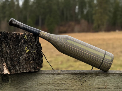 A handmade wood elk bugle with a camo striped grain pattern and carbon pearl acrylic mouthpiece, resting on a weathered wooden fence next to a tree stump with a black lanyard strap.
