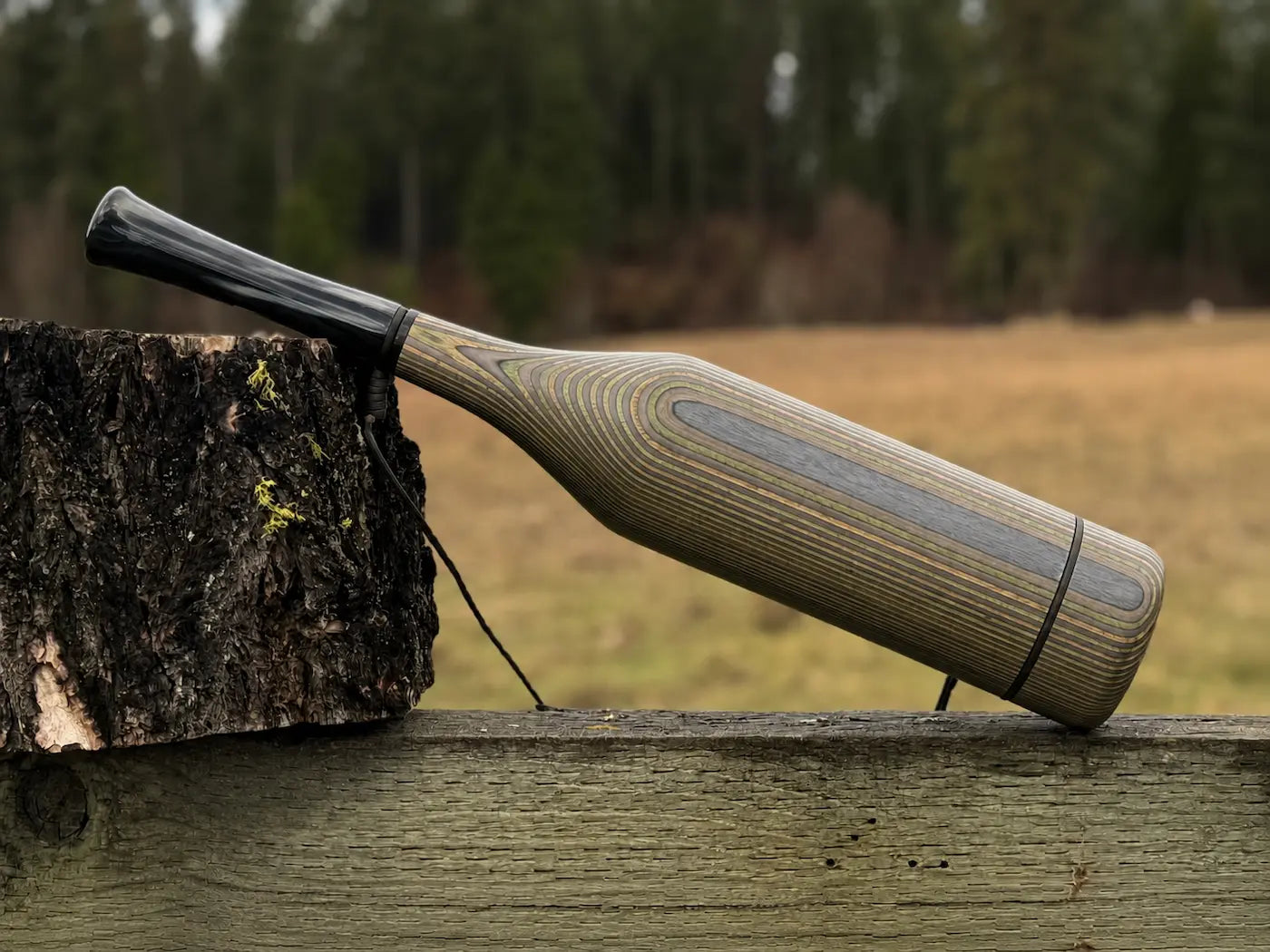 A handmade wood elk bugle with a camo striped grain pattern and black carbon acrylic mouthpiece, resting on a weathered wooden fence next to a tree stump with a black lanyard strap.