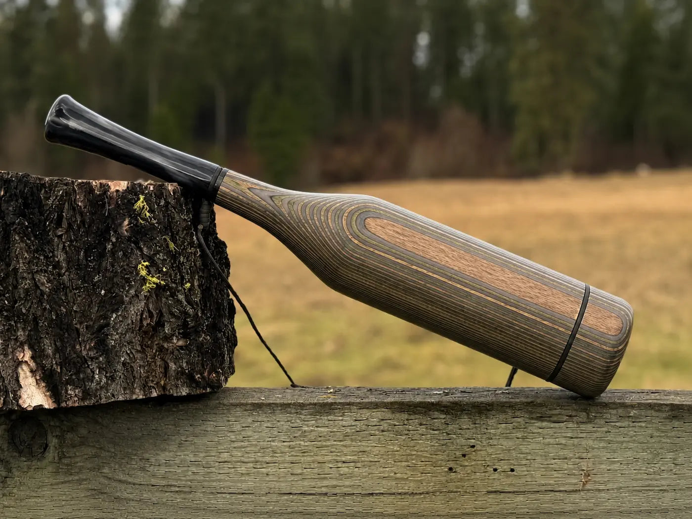 A handmade wood elk bugle with a camo striped grain pattern and carbon pearl acrylic mouthpiece, resting on a weathered wooden fence next to a tree stump with a black lanyard strap.
