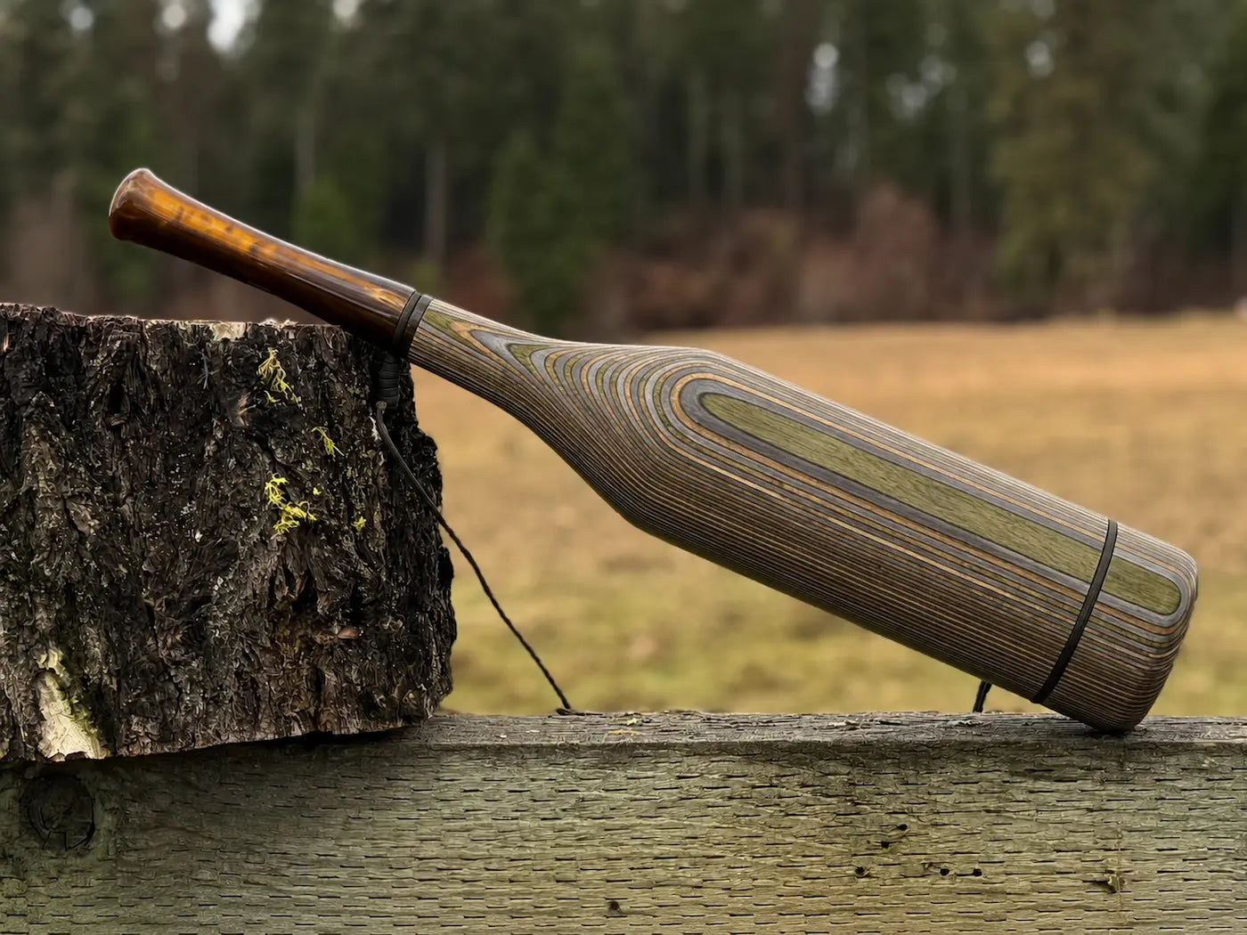 A handmade wood elk bugle with a camo striped grain pattern and bronze pearl acrylic mouthpiece, resting on a weathered wooden fence next to a tree stump with a black lanyard strap.