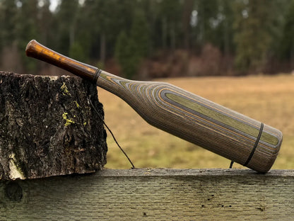 A handmade wood elk bugle with a camo striped grain pattern and bronze pearl acrylic mouthpiece, resting on a weathered wooden fence next to a tree stump with a black lanyard strap.