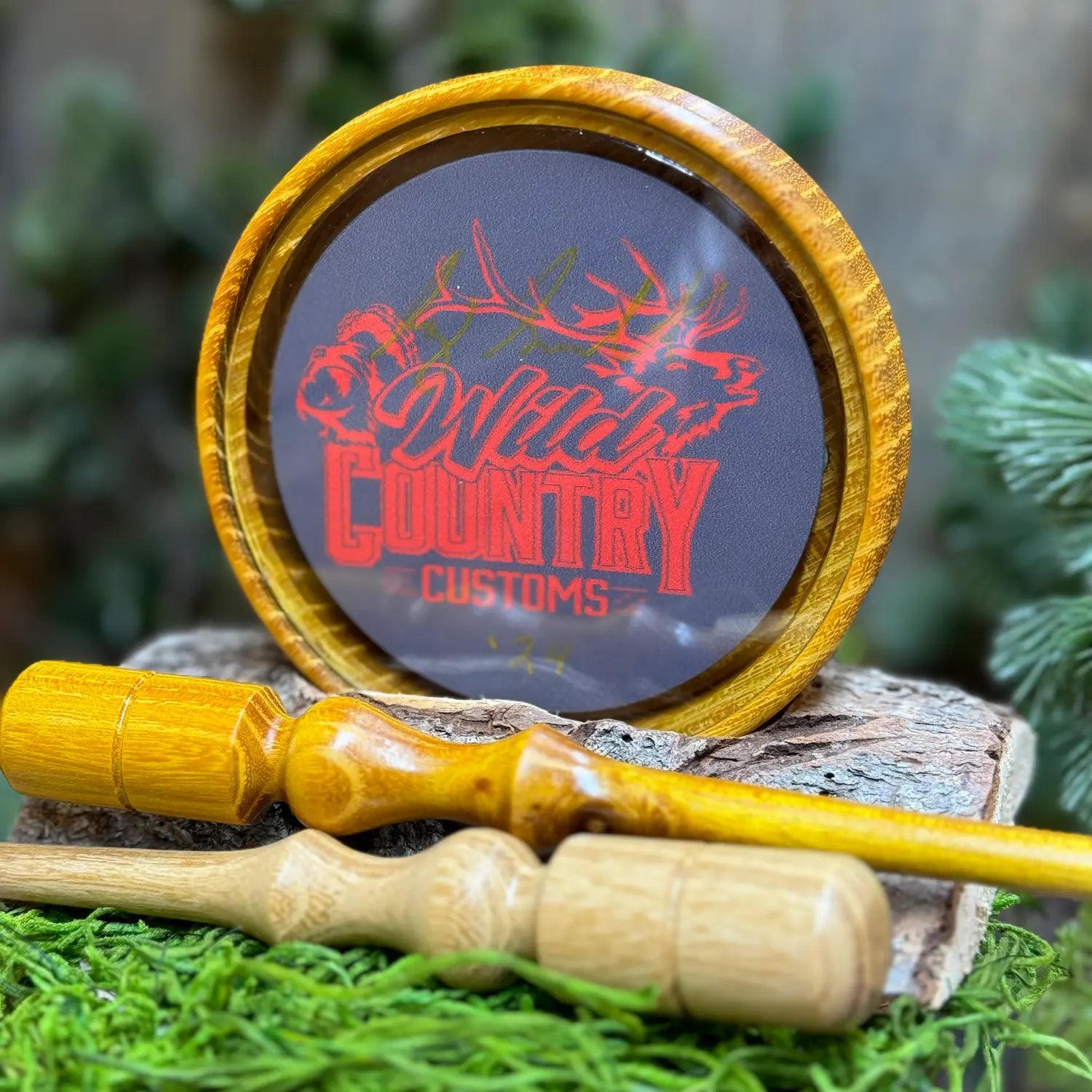Osage Orange Turkey Call with glass playing surface over the Wild Country Customs logo in red. The call is propped up with a matching osage orange striker and a lighter toned black locust wood striker in the foreground