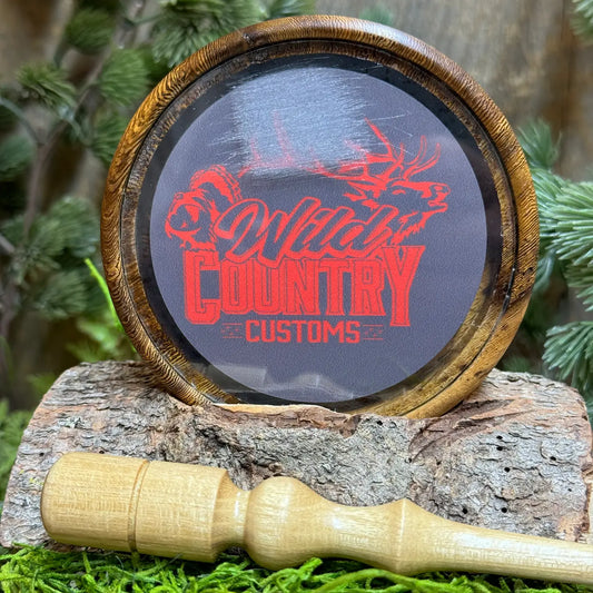 pheasantwood turkey pot call with glass surface and Wild Country Customs logo in red behind the glass. The round pot call is seated upright above a hand crafted light toned black locust wood striker 