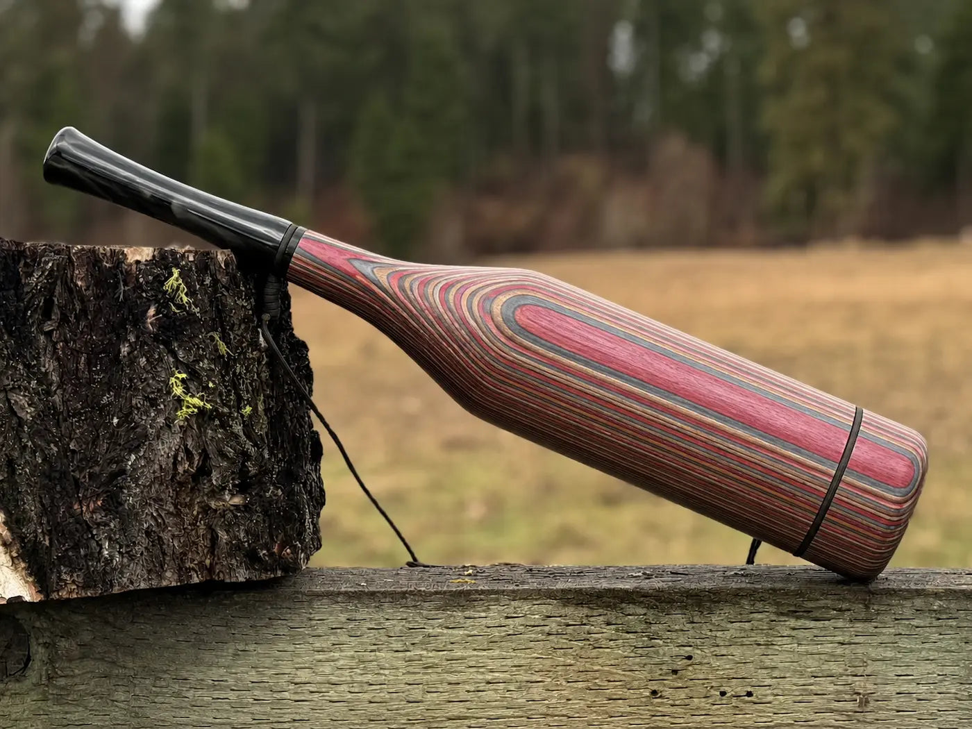 A handmade elk bugle striped multicolor crimson, bronze, and silver laminate wood with a black carbon tone acrylic mouthpiece, resting on a rustic wooden fence next to a tree stump with an outdoor field and forest background.