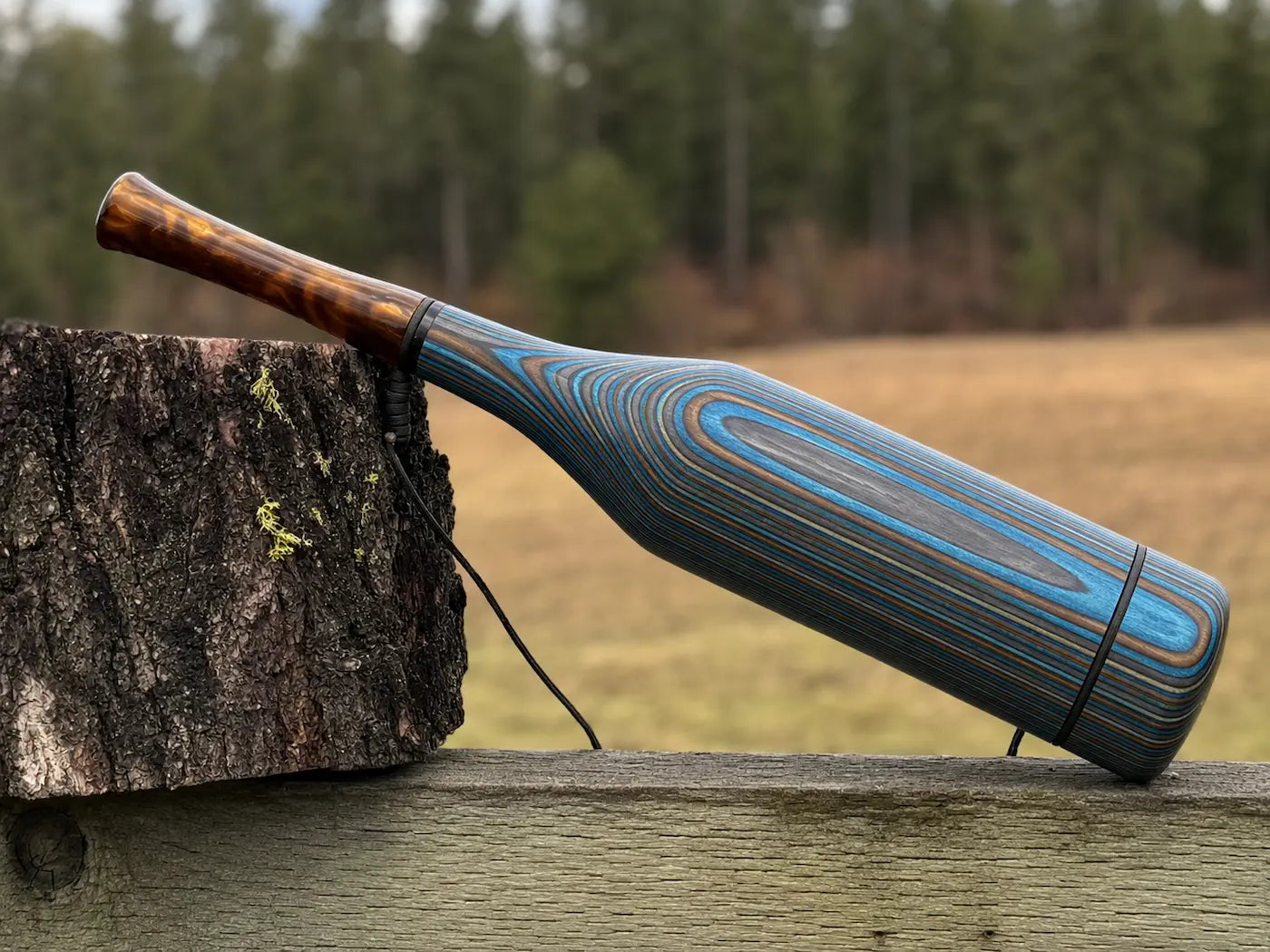 A handmade elk bugle made of blue and bronze striped laminate wood,  having a smoky bronze mouthpiece, rests on a rustic wooden fence next to a tree stump with a blurred forest background.